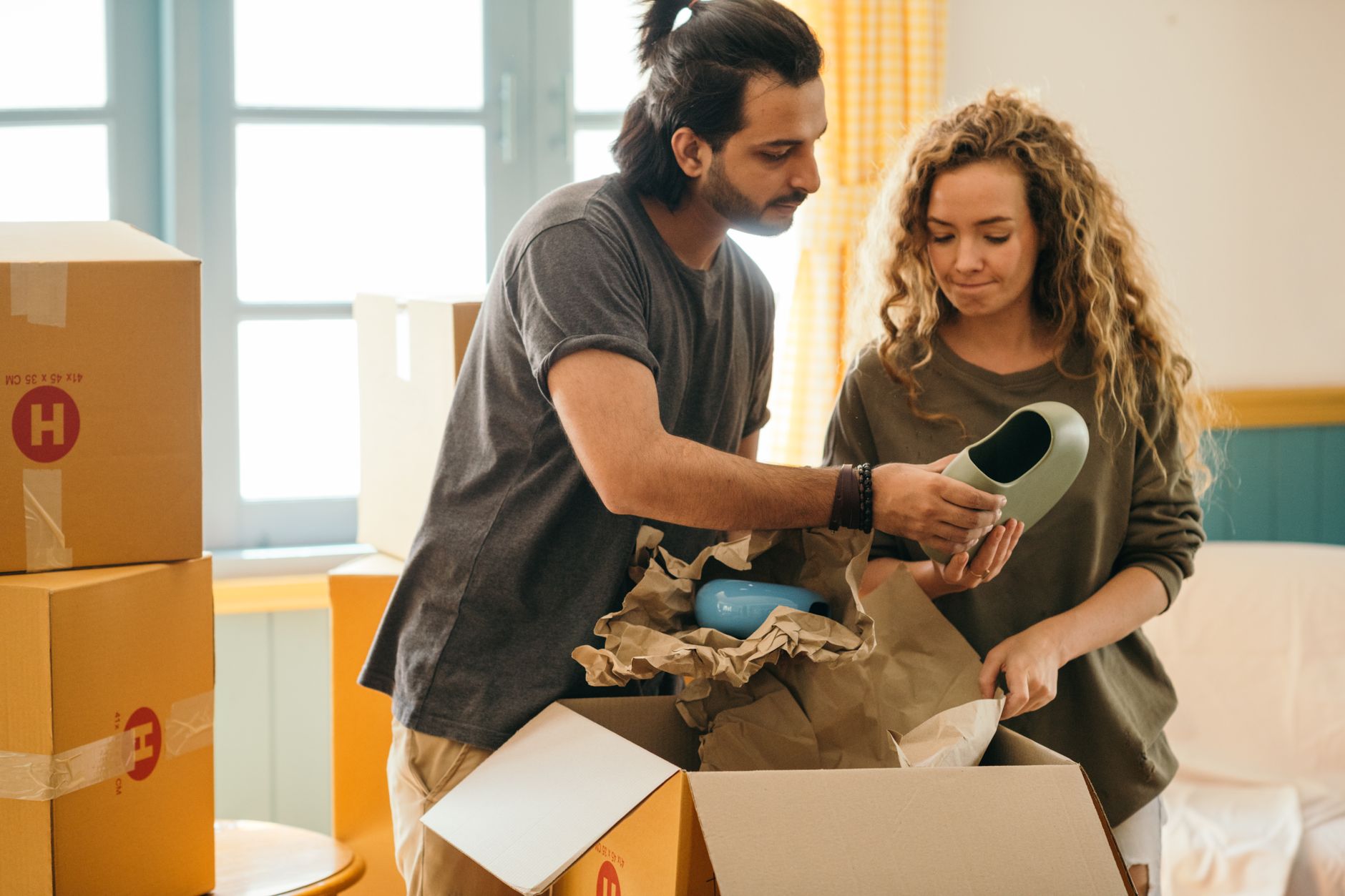 Crop ethnic man showing ceramic vase to girlfriend standing near pile of cardboard boxes with parchment after relocating to n